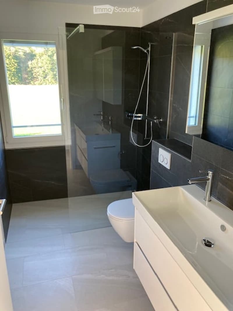 Modern bathroom with dark tiles, white sink, toilet, and shower area. Features a large window and sleek cabinetry.