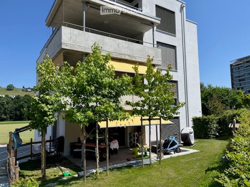 Modern two-story building with a balcony, surrounded by greenery and a small outdoor seating area.