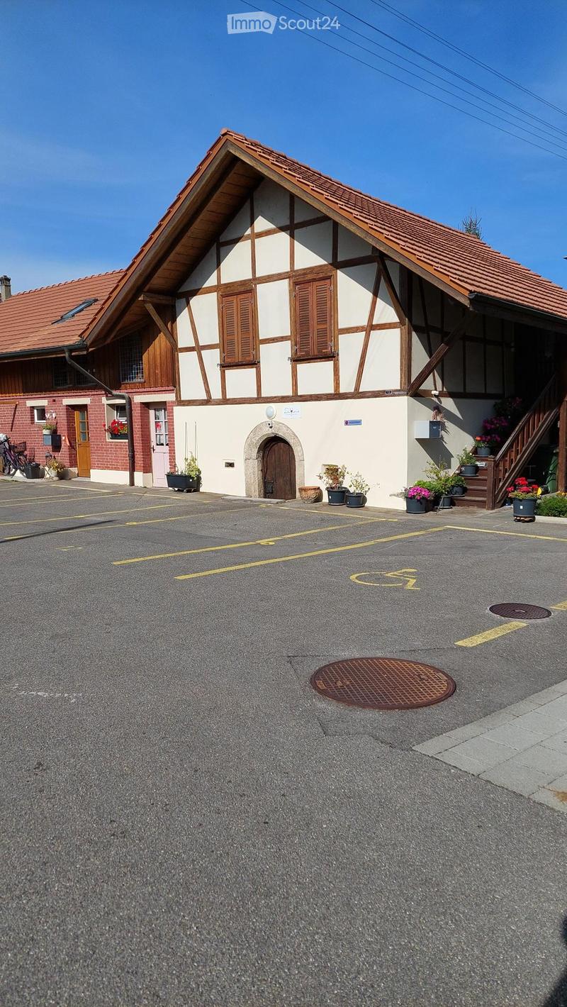 Two-story building with a red-tiled roof and white and wooden facade, featuring multiple windows and doors, surrounded by potted plants and a paved parking area.