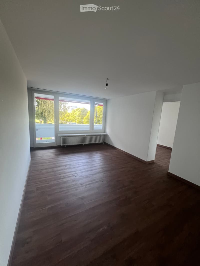 Empty room with wooden flooring, white walls, large windows, radiator, and a visible doorway leading to another space.