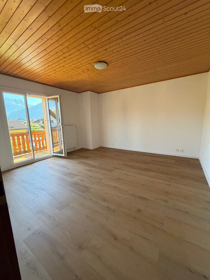 Empty room with wooden flooring, white walls, and a slanted wooden ceiling. A large sliding glass door opens to a balcony with a wooden railing and a scenic mountain view.