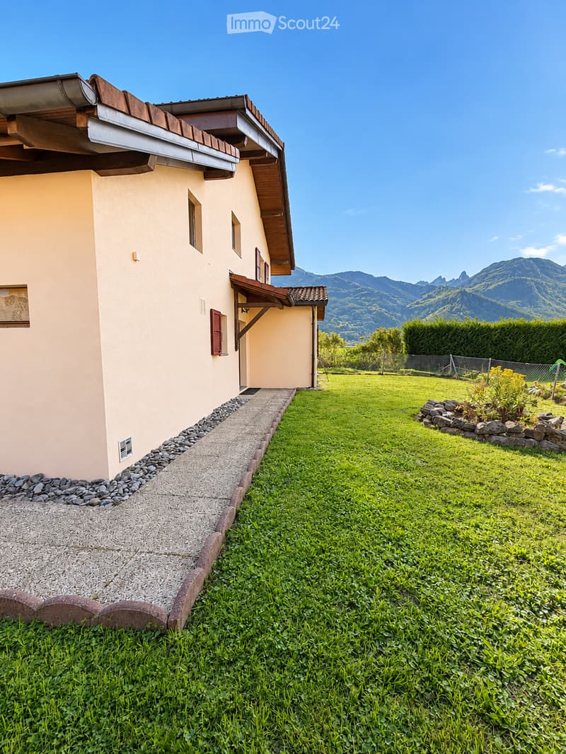 A well-maintained garden area with a gravel pathway leading to a house entrance, surrounded by green grass and a stone border. The house has a light-colored exterior with a wooden roof and a small porch.