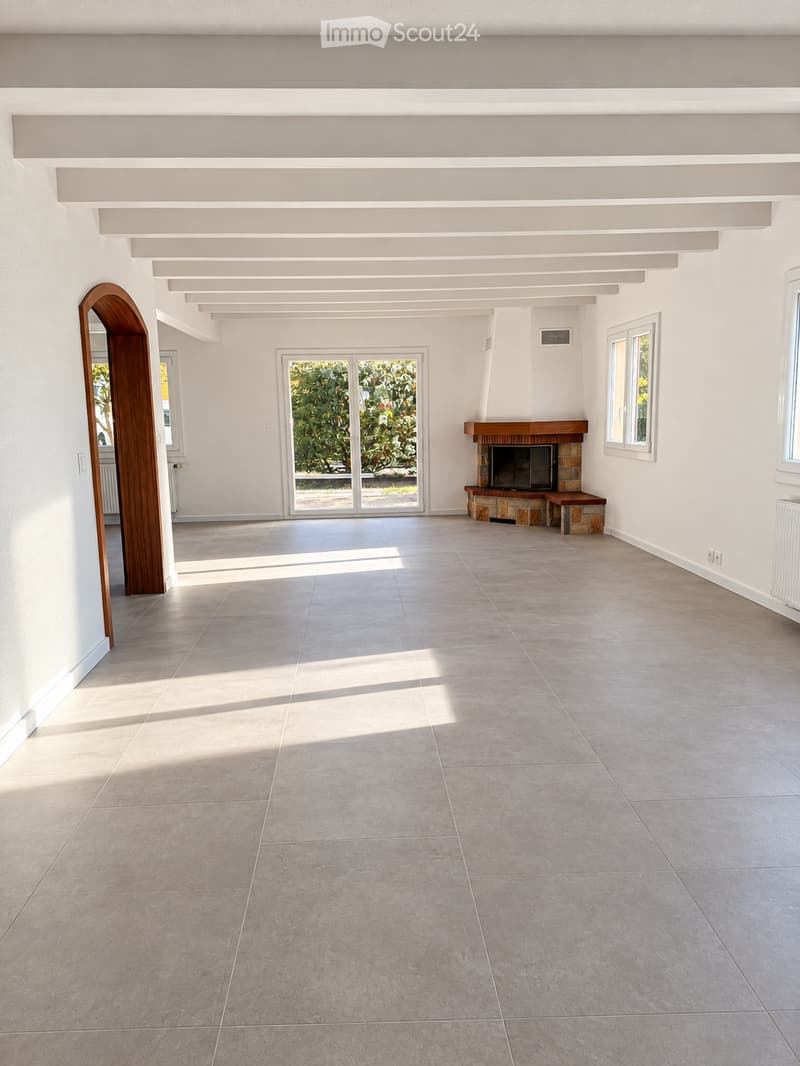 Empty room with tiled floor, white walls, large windows, and a fireplace with a TV on a wooden bench.