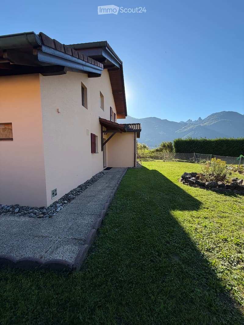 A well-maintained garden area with a gravel pathway leading to a house, surrounded by green grass and a stone border, with a view of mountains in the background.