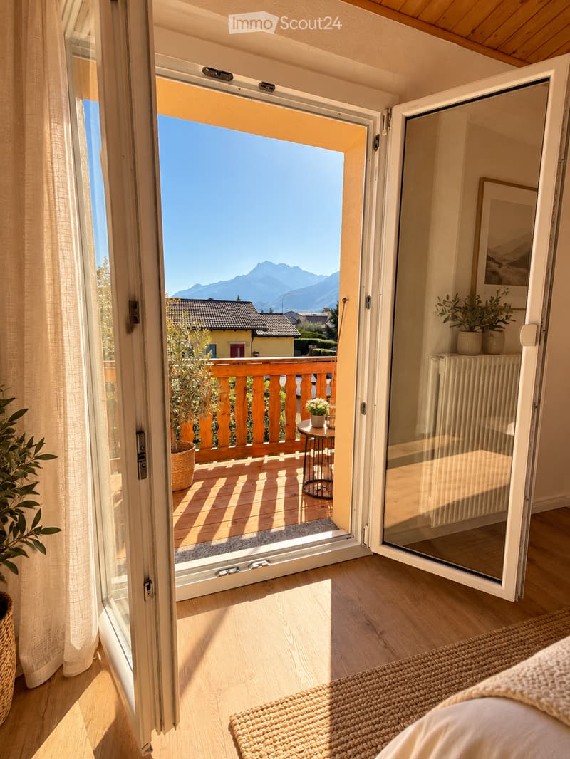 Open sliding glass doors lead to a wooden balcony with a railing, overlooking a scenic mountain view. Inside, a woven rug and potted plants are visible.