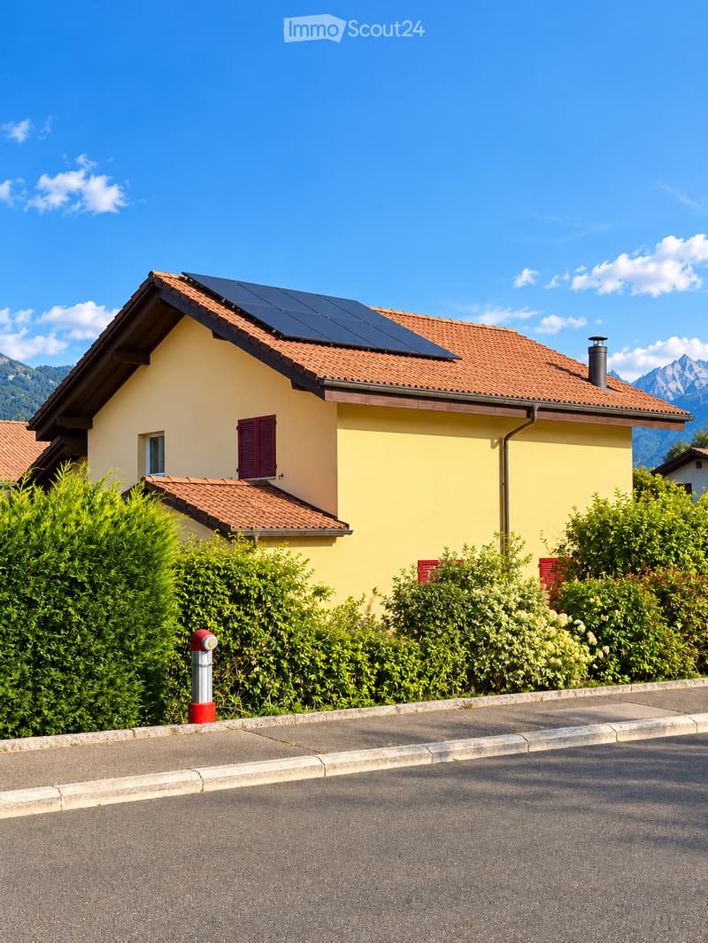 A two-story yellow house with a red-tiled roof, solar panels, and red shutters, surrounded by green bushes and trees, with a clear blue sky and mountain view in the background.