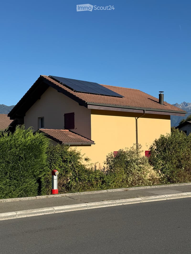Two-story house with a tiled roof, solar panels, and a chimney. The house has light yellow and beige walls, with a small garden area in front.