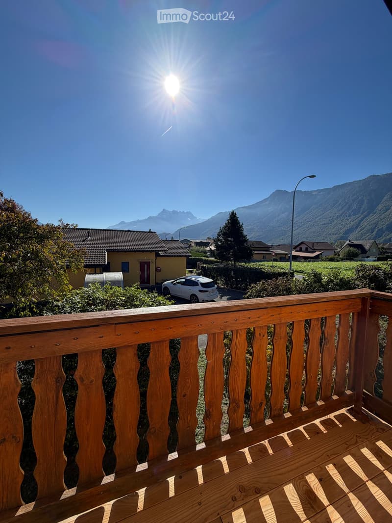 Balcony with wooden railing overlooking a scenic mountain view, with a house and parked car in the foreground.