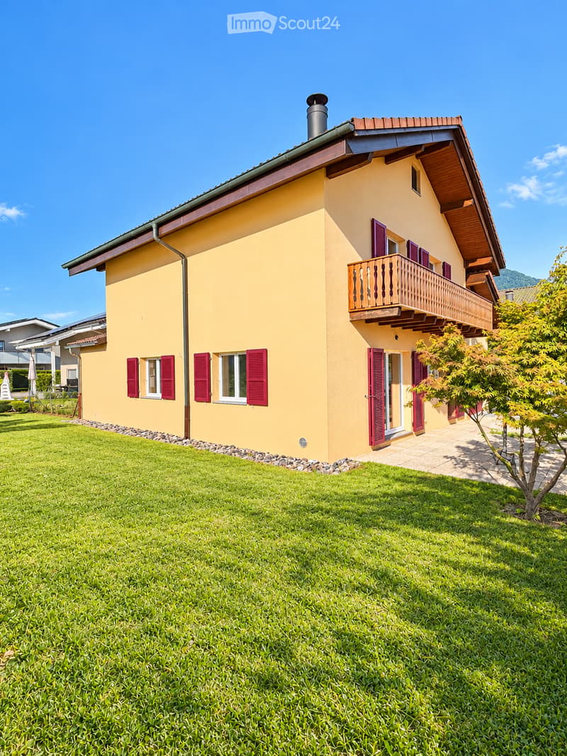 Two-story yellow house with red shutters, a balcony, and a well-maintained lawn in front.