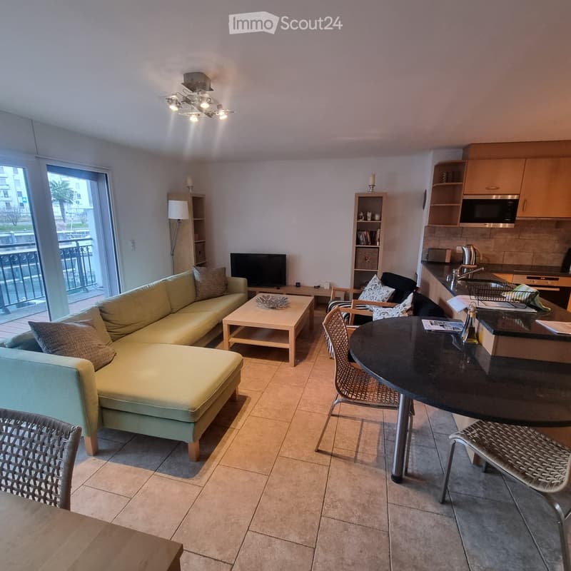 Modern living room with a sofa, coffee table, dining area, and kitchen area. Features a balcony with outdoor view.