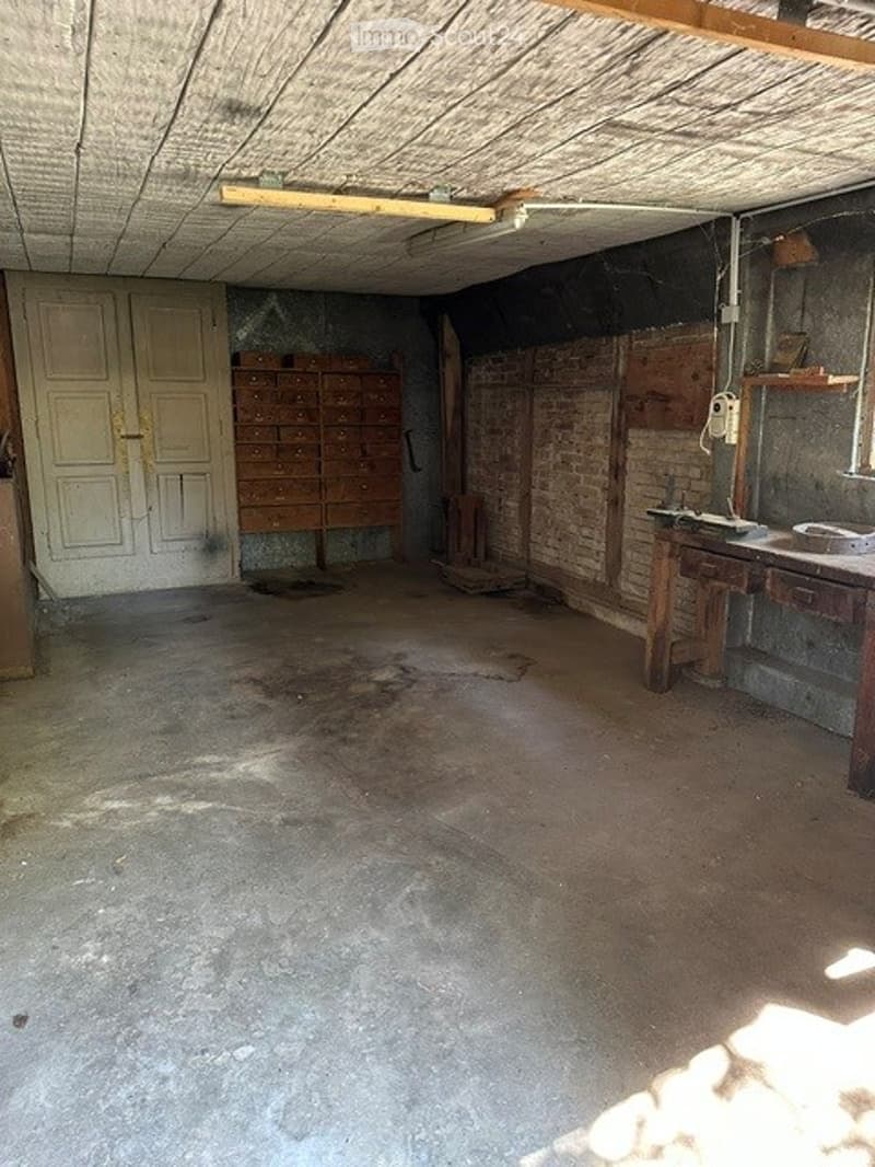 Empty room with exposed brick walls, wooden ceiling beams, and a concrete floor. Features include a wooden filing cabinet, a small wooden desk, and a white door.