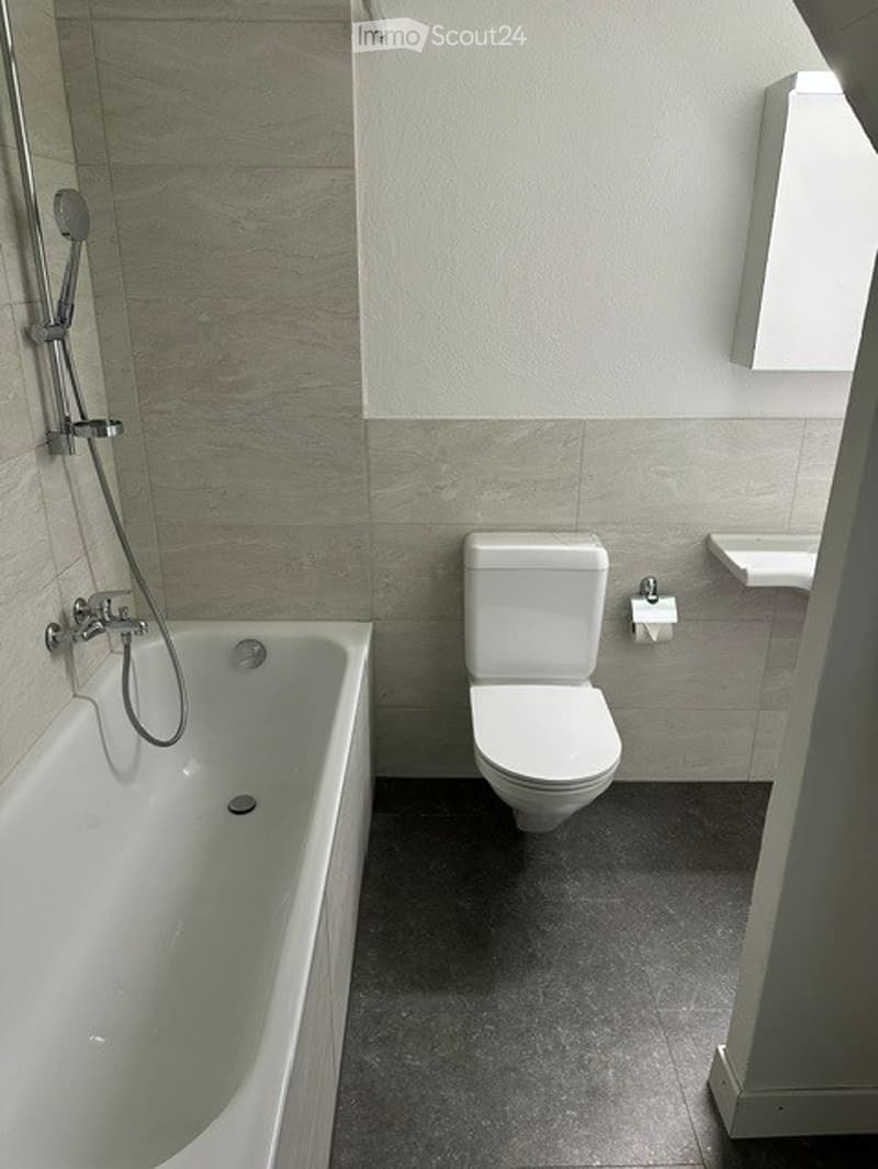 Modern bathroom with a bathtub, toilet, sink, and showerhead. The walls are tiled in a neutral color scheme, and the floor is dark grey.