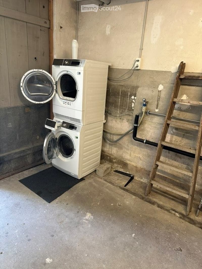 Utility room with a stacked washing machine and dryer, concrete floor, exposed pipes, and a wooden ladder.