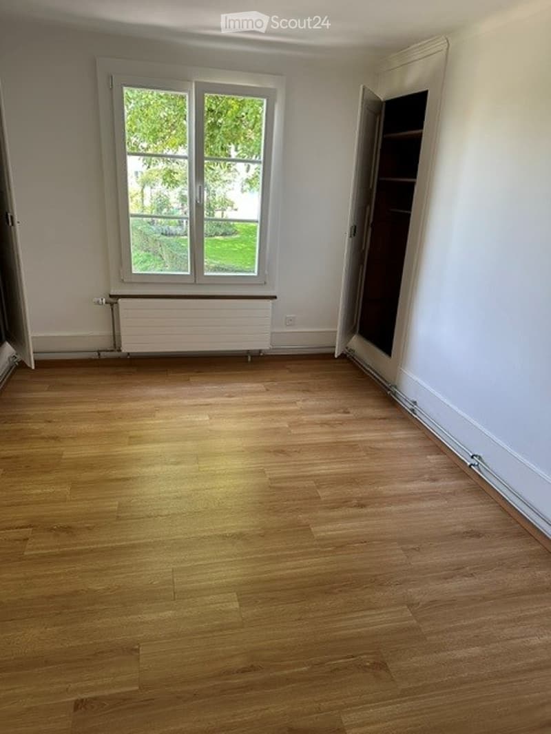 Empty room with wooden flooring, white walls, a large window with a view of greenery, and a radiator below the window.