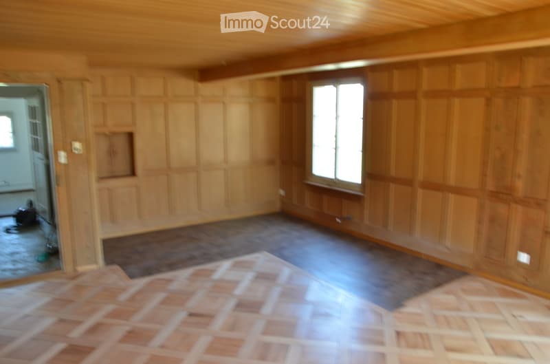 Empty room with wooden panel walls, a mix of wooden and tiled flooring, and large windows allowing natural light.
