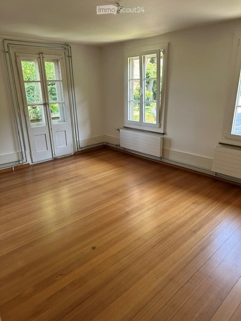 Empty room with wooden flooring, white walls, and large windows with white frames. No furniture or equipment present.