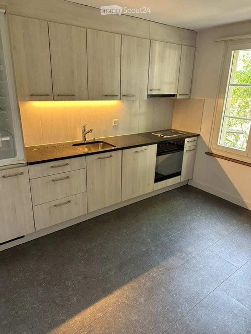 Modern kitchen with light wood cabinets, stainless steel sink, and built-in oven. The space features a dark countertop and tiled floor.