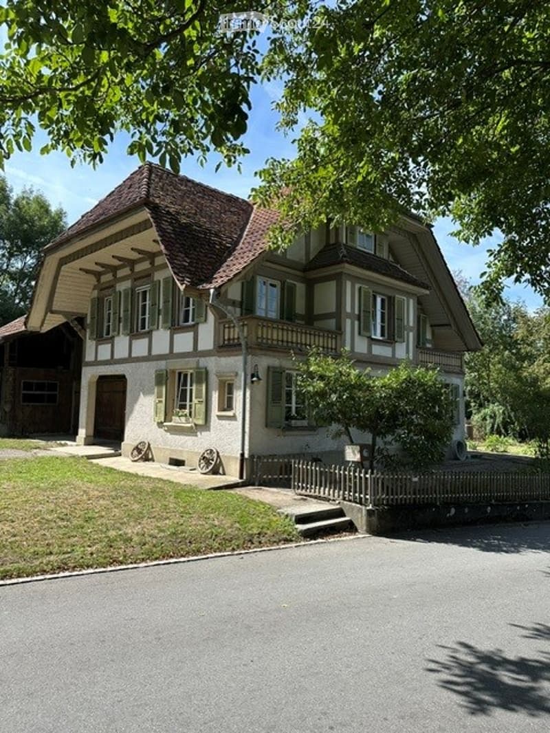 Two-story house with a gabled roof, white siding, green shutters, and a small balcony on the upper level. The house is surrounded by a wooden fence and trees.