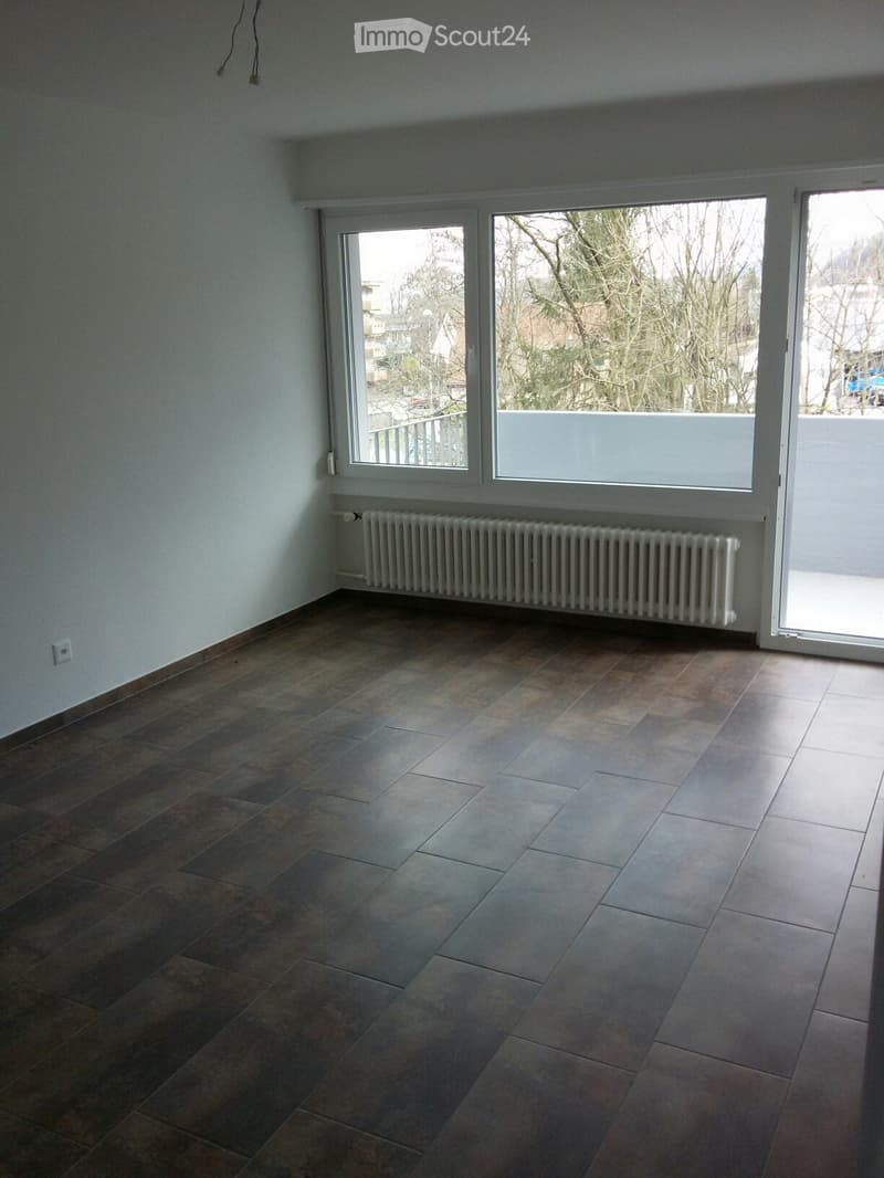Empty room with tiled floor, white walls, large windows, radiator, and sliding glass door leading to a balcony.