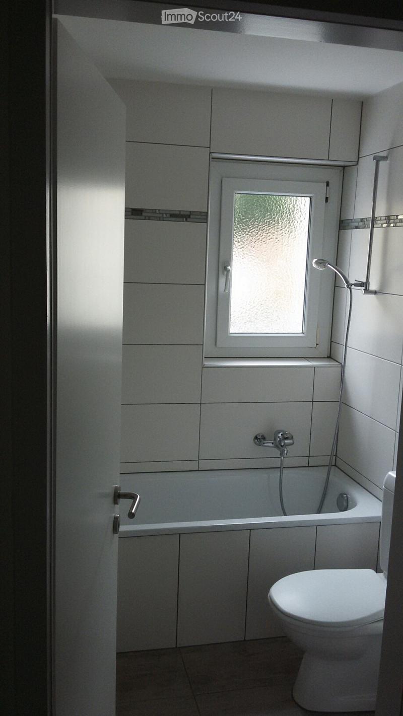 Modern bathroom with white tiled walls, a bathtub, toilet, and a showerhead attached to the wall. A frosted window provides natural light.