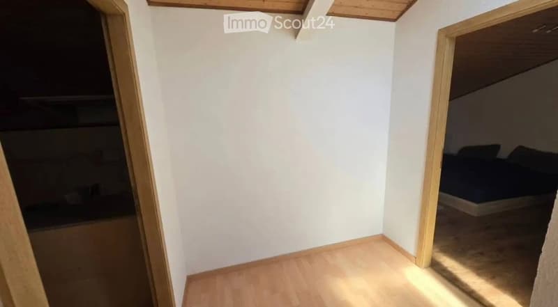 Empty room with light wooden flooring and white walls, featuring an open doorway leading to another room with a bed visible in the background.