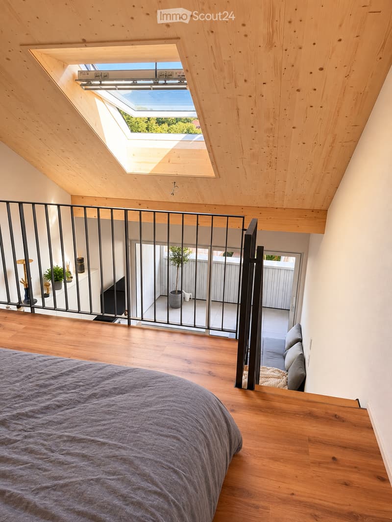 Modern bedroom with wooden floors, a large skylight, and a balcony accessible through a black metal railing. The room features a grey bedspread, a small table with decorative items, and a potted plant on the balcony.