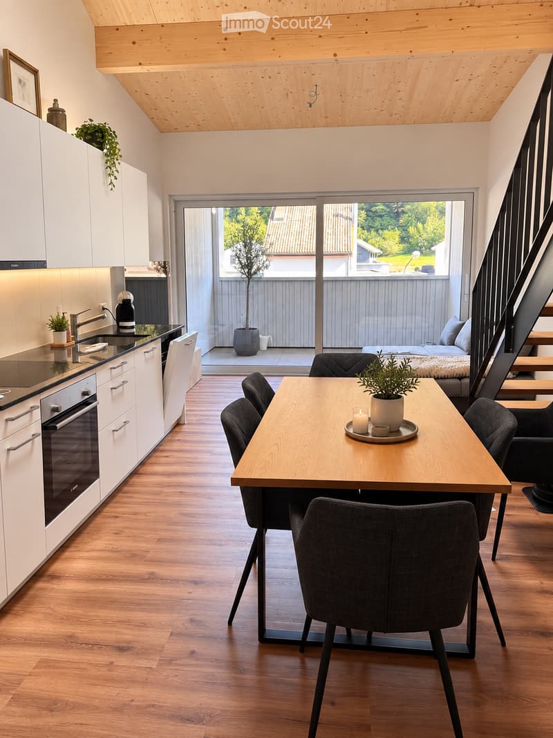 Open-plan living and dining area with modern white cabinets, a wooden dining table with chairs, and a kitchen area featuring an oven, sink, and faucet. A staircase leads to an upper level with a bed visible.