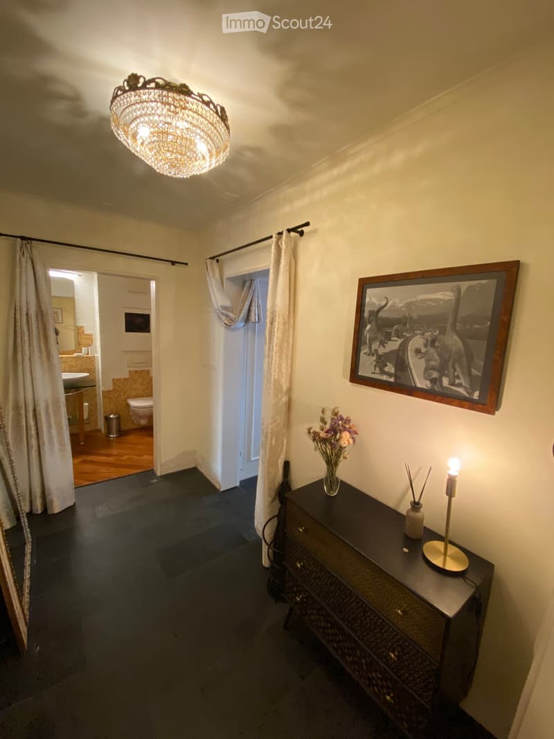 Hallway with dark flooring, white walls, a chandelier, a framed picture, a small table with a vase of flowers, and a lit candle. An open doorway leads to a bathroom with wooden flooring and a toilet.
