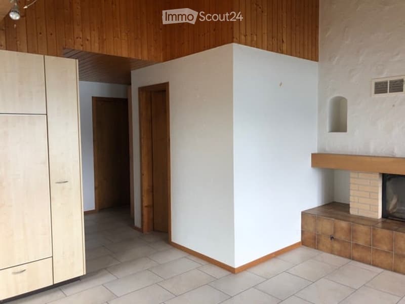 Empty room with white walls, wooden ceiling, tiled floor, and a fireplace with a wooden bench. There are built-in wooden cabinets and a doorway leading to another room.