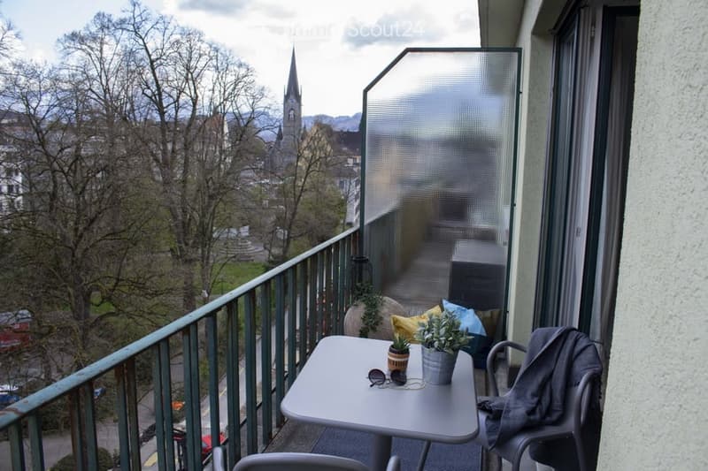 Balcony with a view of a town and mountains, featuring a small table, chairs, and potted plants.