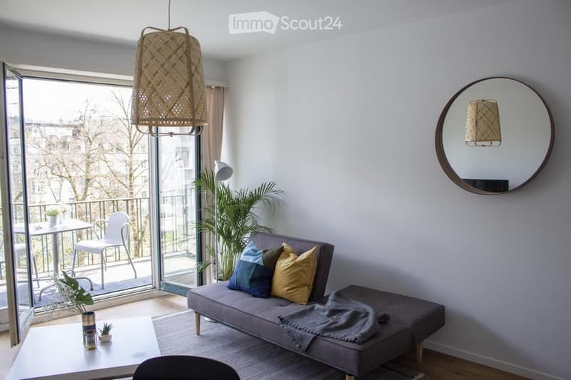 Modern living room with a grey sofa, colorful pillows, a coffee table, a round mirror, and a balcony with outdoor furniture.