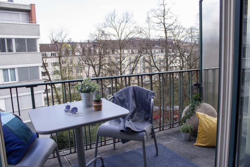 Balcony with a small table, chairs, and potted plants, overlooking a cityscape with buildings and bare trees.
