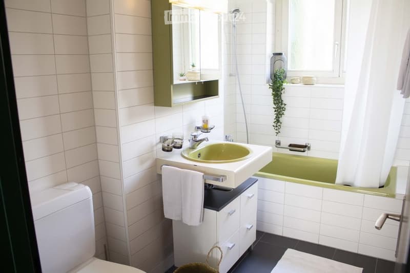 Modern bathroom with white tiled walls, a green sink, a bathtub with a shower, a mirror cabinet, and a towel on the cabinet.