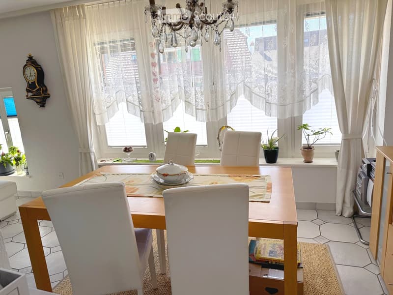 Dining room with a wooden dining table, six white chairs, a chandelier, a clock on the wall, and potted plants on the windowsill.