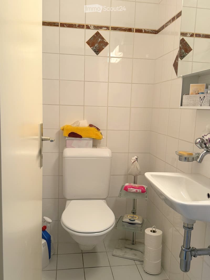 A small bathroom with white tiles, a toilet, a sink, a mirror, a glass shelf with soap and other items, and a towel on top of the toilet tank.