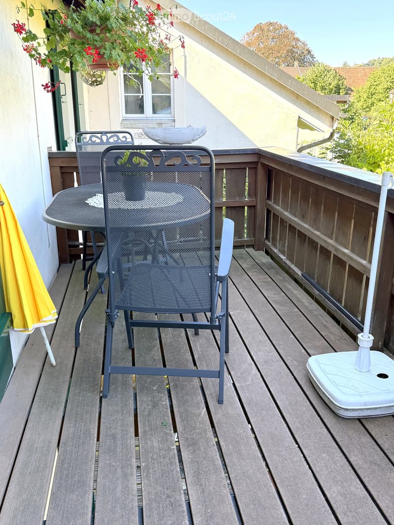 Wooden balcony with a metal chair, small table, and umbrella stand. Potted plants and a decorative bowl are visible in the background.