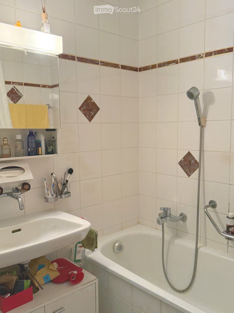 Bathroom with white tiles, a sink, bathtub, mirror, and various toiletries. Features a showerhead and a towel on the edge of the tub.