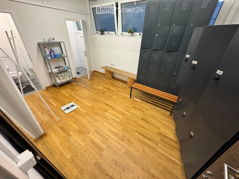 The room features wooden flooring, several lockers, a small bench, a shelf with cleaning supplies, a toilet, and a scale on the floor.