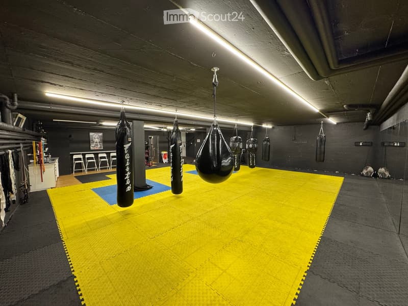 A spacious gym area with a yellow and blue padded mat, several hanging boxing bags, a few chairs, and a small cabinet with various items.