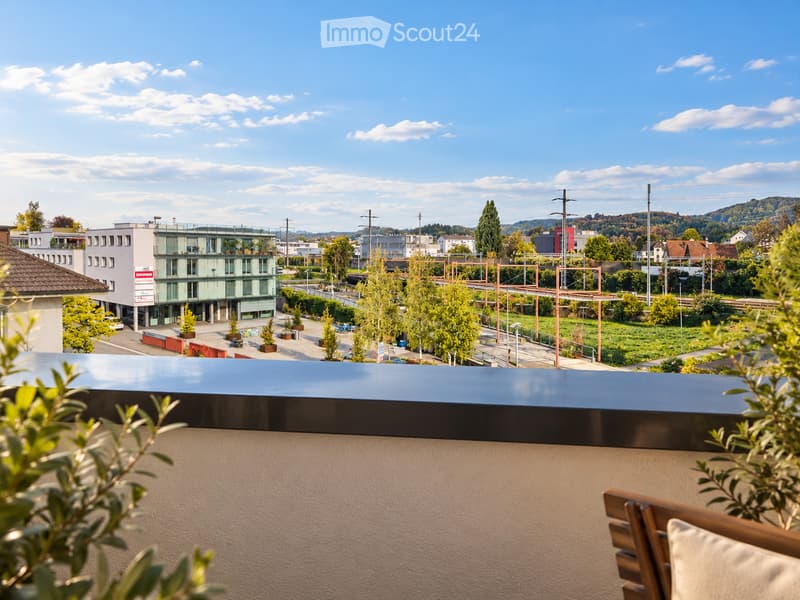 Balcony with a wooden chair and a view of a cityscape, including buildings, trees, and a clear blue sky.