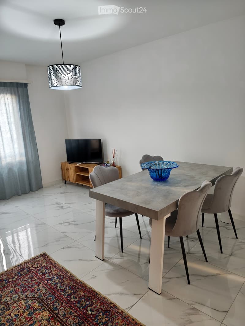 Modern dining area with a marble floor, a large dining table with four chairs, a pendant light, a television on a wooden stand, and a decorative rug.