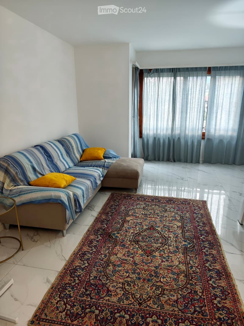Living room with a sofa, ottoman, large patterned rug, and a small round glass table.