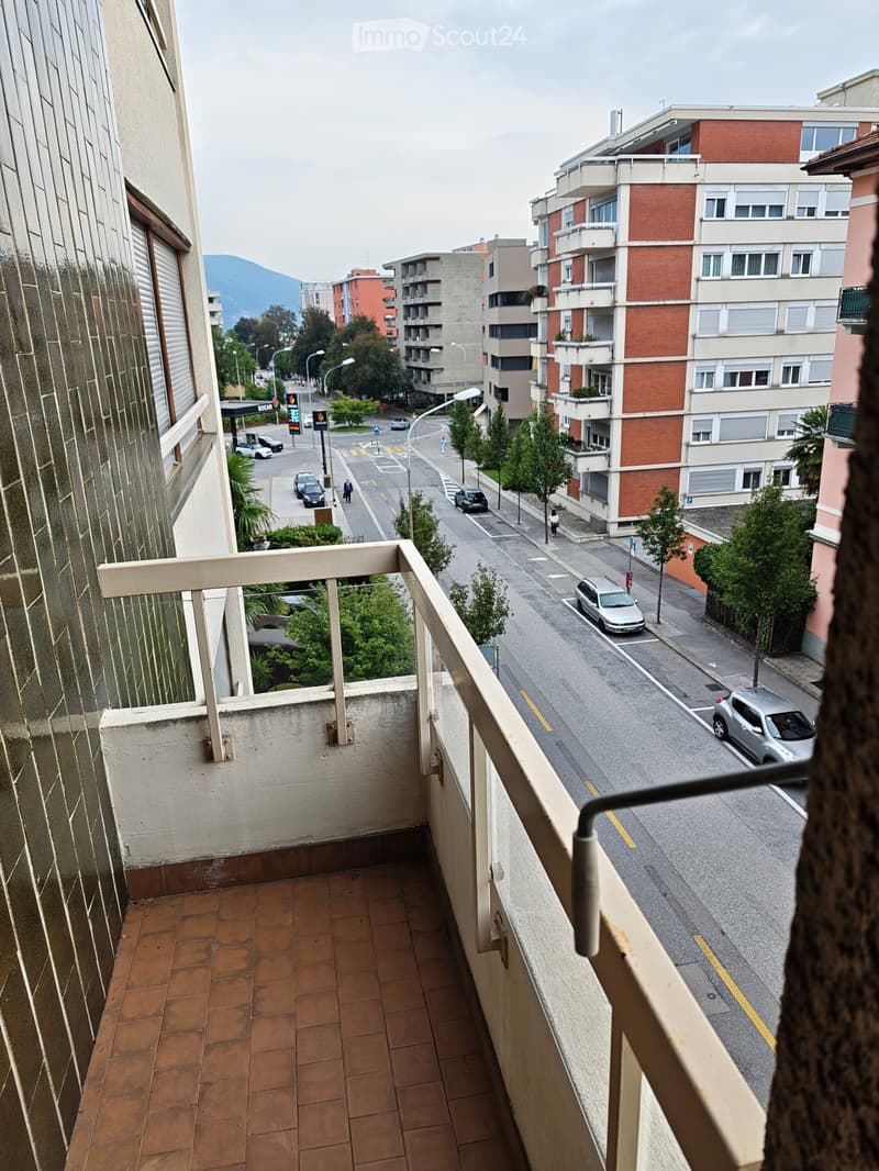 Balcony with tiled floor and wooden railing overlooking a street lined with parked cars and buildings.