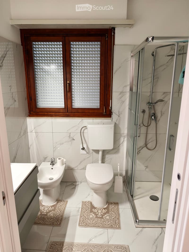 Modern bathroom with marble tiles, a toilet, bidet, shower, and a window with frosted glass.