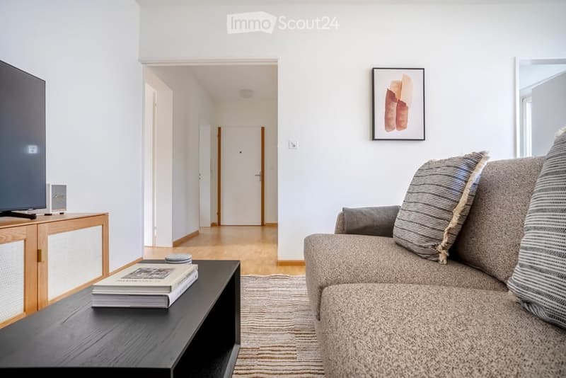 Modern living room with a grey sofa, coffee table, books, and a TV stand. The space features light-colored walls, wooden flooring, and a hallway leading to another room.