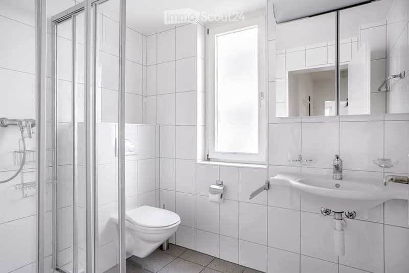 Modern bathroom with white tiled walls and floor, featuring a toilet, sink with mirror, and a glass-enclosed shower area.