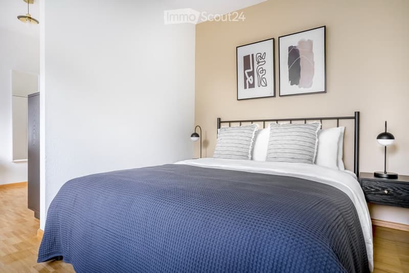Modern bedroom with a neatly made bed, nightstand with lamp, and two framed artworks on the wall. Light-colored walls and wooden flooring.