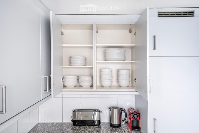 Modern kitchen with white cabinets, open shelving displaying plates, and countertop appliances including a toaster, kettle, and coffee maker.