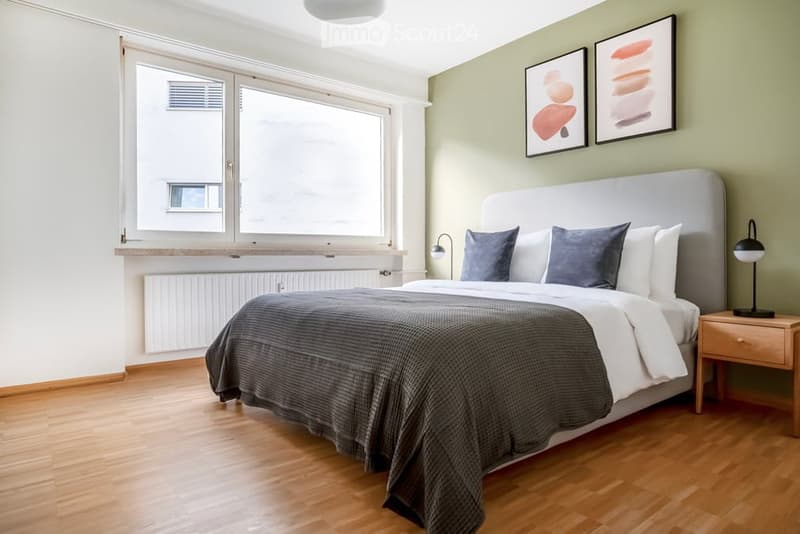 Modern bedroom with a large bed, nightstand, lamp, and two picture frames on the wall. The room features wooden flooring and large windows.
