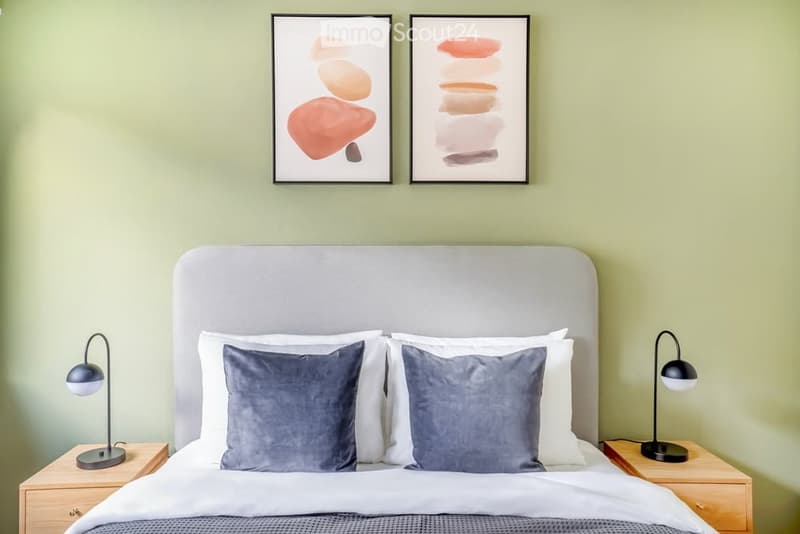 A modern bedroom with a large bed, two nightstands, lamps, and decorative pillows. The walls are painted a soft green, and there are two framed abstract art pieces above the bed.